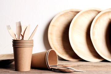 Eco-friendly tableware on wooden table and white wall. Bamboo plates, paper cups and wooden cutlery.