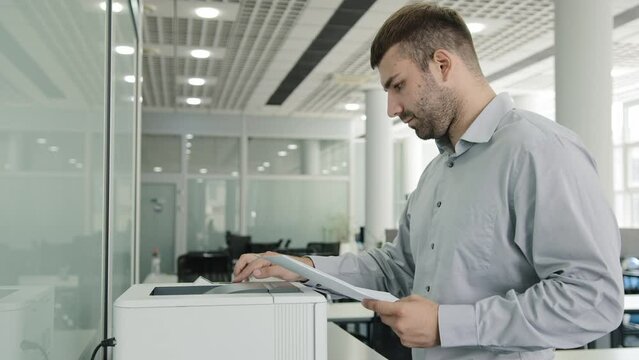 Focused caucasian male professional worker manager prints documents on office equipment makes copy on copier uses printer replicate papers using electronic print machine presses start button printing