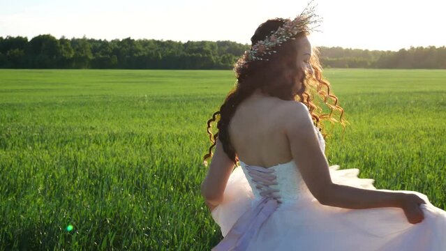 Bride is spinning in a wedding dress in wheat field against the backdrop of forest and she smiles. Wedding dress sways in the wind. Bride has wreath on her head.