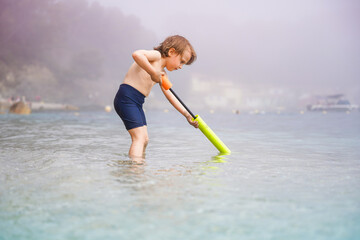 Little toddler boy playing with water toy blaster in the sea in foggy cloudy weather. Child spending summer vacation at the seaside in Europe.