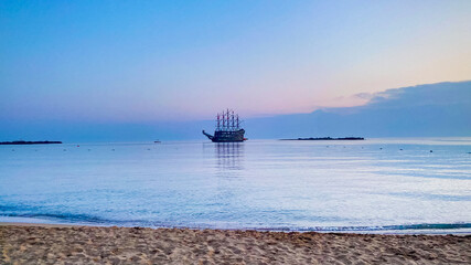 An old pirate ship is anchored in the sea near the sandy beach at sunset