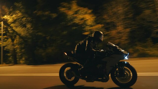 Young man in helmet riding fast on modern sport motorbike at evening city street. Motorcyclist racing his motorcycle on night empty road. Guy driving bike. Concept of freedom and hobby. Side view