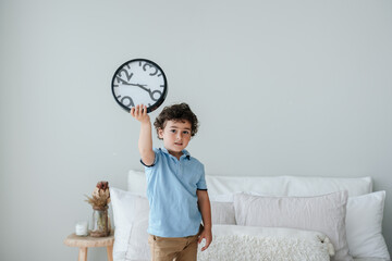 Cute caucasian boy in blue polo holding wall clock standing on bad, early morning getting up. Time...