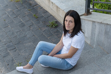 A young brunette woman in a white t-shirt and blue jeans sits on the stairs in the park.