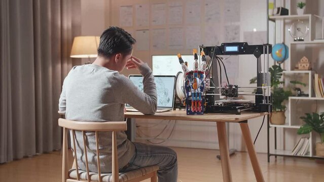 Back View Of A Male With 3D Printing Having A Headache While Designing A Cyborg Hand On A Laptop At Home
