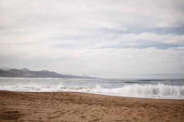 Ocean waves with splashes on the beach sand with mountains on the background on a cloudy day with grey clouds