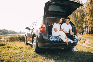 Happy traveler couple sitting in car open trunk and watch sunrise