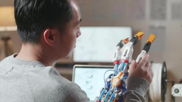 Close Up Side View Of Asian Male With 3D Printing Checking A Cyborg Hand While Designing It On A Laptop At Home

