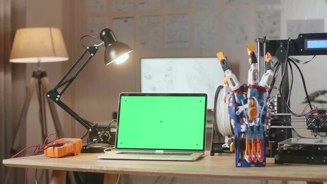 A Laptop With Mock-Up Green Screen Is Next To A Cyborg Hand And 3D Printing On The Table At Home
