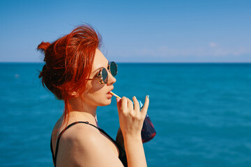 Red-haired woman in sunglasses drinks lemonade from bottle through cocktail tube. Girl on summer vacation. Blue sea surface and clear sky on background. Copy space for travel agency advertising.