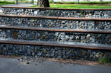 grandstand in the park by a concrete staircase made of gabion baskets with gray stones arranged inside. the seating areas of the benches are steps made of planks.