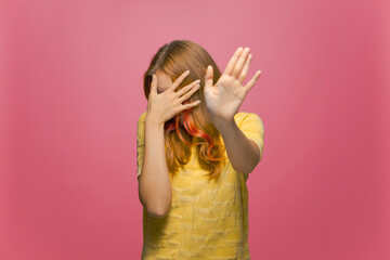 Scared or embarrassed young girl covering eyes with hands and doing stop gesture on pink studio...