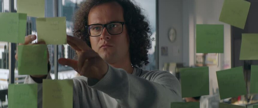 Young Caucasian male employee looking at adhesive sticky notes to a glass wall in the modern office, scrum agile methodology planning project. Shot through the glass