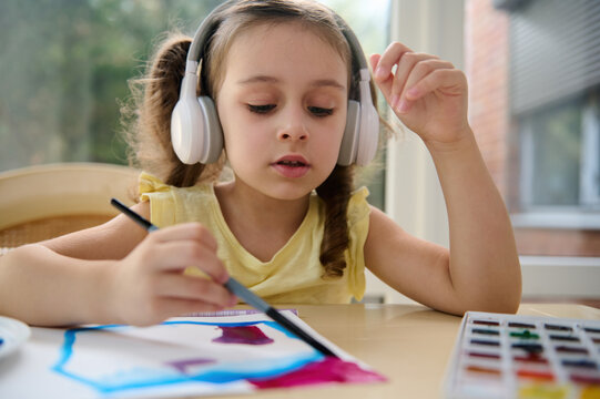Close-up portrait of adorable European preschool child, cute baby girl in audio headset, painting picture with brush and watercolor. Creativity courses for children, talented little artists, painters