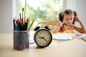 Focus on black alarm clock and pencil holder on the blurred background of little girl of preschool age, sitting at table and drawing picture with watercolors. Painting. Art class and courses for kids - Powered by Adobe