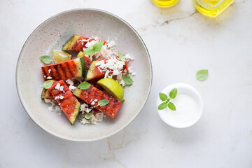 Grilled watermelon with grated cheese and yogurt, flat lay on a light-beige marble background, horizontal shot