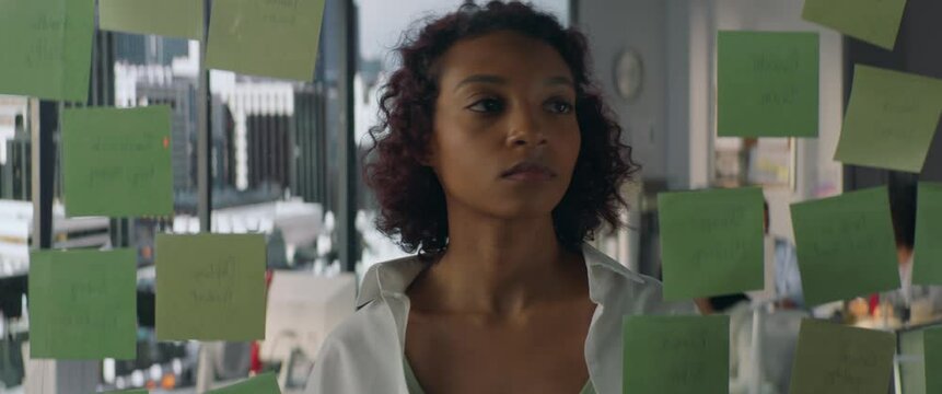 Young African American female employee looking at adhesive sticky notes on glass in the modern office, scrum agile methodology planning project. Shot through the glass