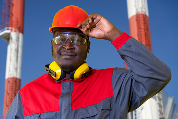 African American Worker In Protective Workwear Standing In Front Of Industrial Chimneys. Portrait Of Black Power Plant Worker In Red Helmet, Protective Eyewear And Hearing Protection Equipment.