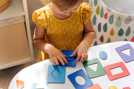 A little girl playing with wooden shape sorter toy on the table in playroom. Educational boards for Color and Shapes sorting for toddler. Learning through play. Developing Montessori activities.