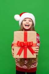 A little girl holds a gift and holds it forward on a green isolated background. A child in a Santa Claus hat and a warm sweater holds a gift box.