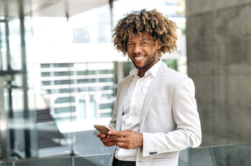 Handsome happy brazilian or hispanic man with curly hair, stylishly elegantly dressed, standing outdoors near the business center, using mobile phone, online messaging, looks at camera, smiling