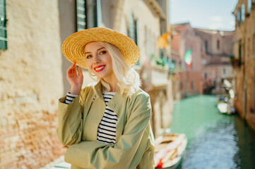 Happy smiling fashionable woman wearing straw hat, striped t-shirt, green trench coat, posing on the bridge in Venice, Italy. Fashion, travel, vacation, lifestyle concept. Copy, empty space for text