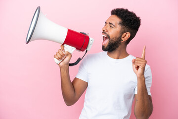 Young Brazilian man isolated on pink background shouting through a megaphone to announce something in lateral position