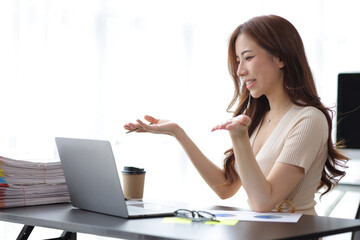 Beautiful Asian businesswoman sitting in her private office, she is talking to her partner via video call on her laptop, she is a female executive of a startup. Concept of financial management.