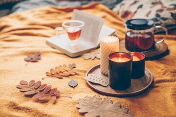 Autumn cozy at home. Burning candles with card hello autumn, herbal tea in glass cup and open book on yellow plaid.