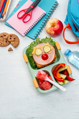 School lunch box for kids with food in the form of funny faces. School lunch box with sandwich, vegetables, water and stationery on table. Healthy eating habits concept. Back to school concept