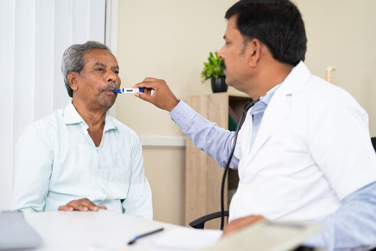 Doctor at clinic checking temperature of sick old man using thermometer due to fever - concept of medical treatment, counselling and health checkup