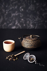 Golden teapot, bowl, infuser and bamboo spoon with tea strands on black surface and textured gray background, space for advertising. 