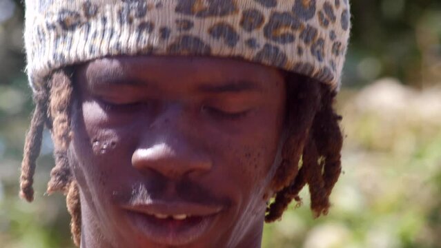 Footage of black man head with hat and dreadlocks coming out of it concentrating at playing dundun or dunun drum, from the front.