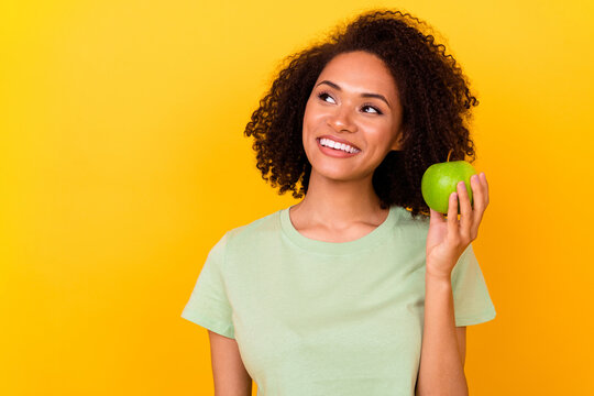 Photo of positive dreamy lady wear green t-shirt eating apple white teeth looking empty space isolated yellow color background
