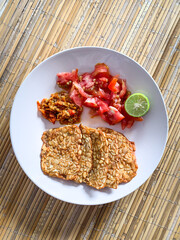 fried tempeh with slices tomato and chilli sauce on a white plate.