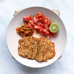 fried tempeh with slices tomato and chilli sauce on a white plate.