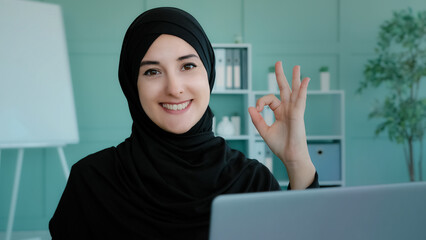 Muslim businesswoman freelancer in black hijab working typing on laptop using computer service for shopping online Islamic girl student studying shows ok gesture approves agree recommendation sign