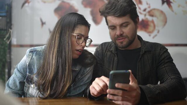 Couple looking at smartphone screen seated at coffee shop. People laughing and smiling over video content online. Young man showing phone screen to girl