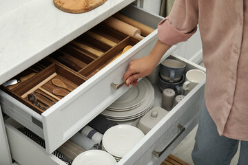Woman opening drawers of kitchen cabinet with different dishware and utensils, closeup