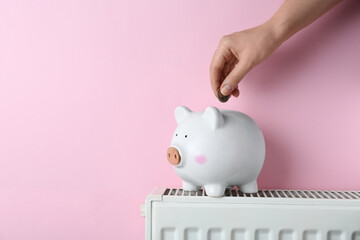 Woman putting coin into piggy bank on heating radiator against pink background, closeup. Space for text