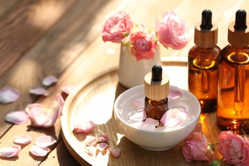 Bottles of rose essential oil and flowers on wooden table, space for text