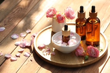 Bottles of rose essential oil and flowers on wooden table, space for text