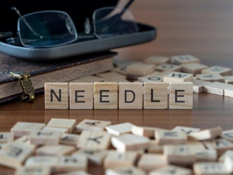 needle word or concept represented by wooden letter tiles on a wooden table with glasses and a book