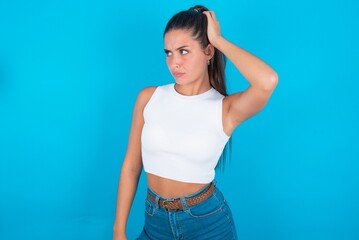 beautiful brunette woman wearing white tank top over blue background confuse and wonder about question. Uncertain with doubt, thinking with hand on head. Pensive concept.