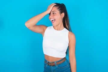 beautiful brunette woman wearing white tank top over blue background surprised with hand on head...
