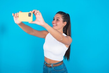 beautiful brunette woman wearing white tank top over blue background taking a selfie to post it on social media or having a video call with friends.