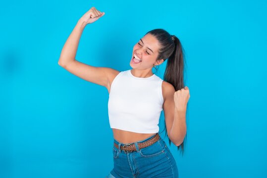 Profile photo of excited beautiful brunette woman wearing white tank top over blue background good mood raise fists screaming rejoicing overjoyed basketball sports fan supporter
