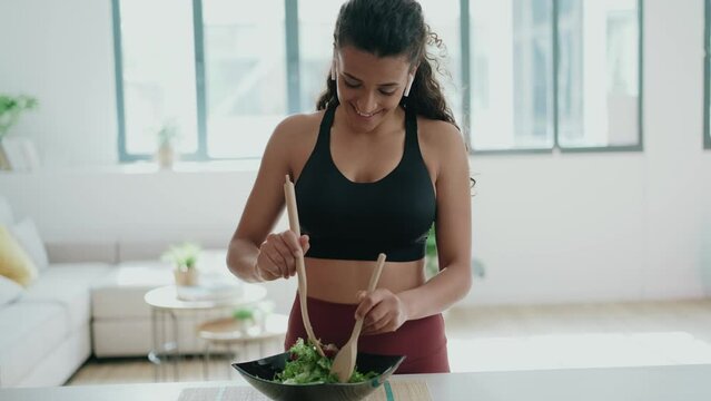 Video of beautiful young woman making a healthy salad while singing and listening music with headphones in the kitchen at home.