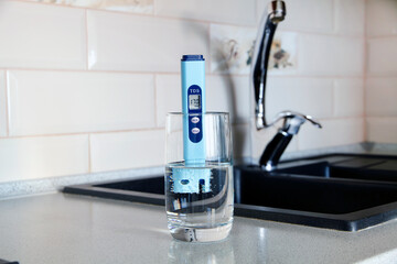  Electronic pH meter in a glass of water. In the background there is a tap for drinking water. The...