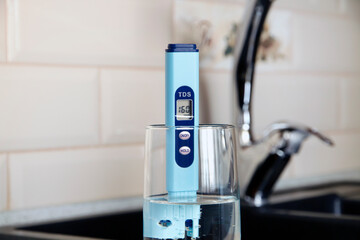 Electronic pH meter in a glass of water. In the background there is a tap for drinking water. The...